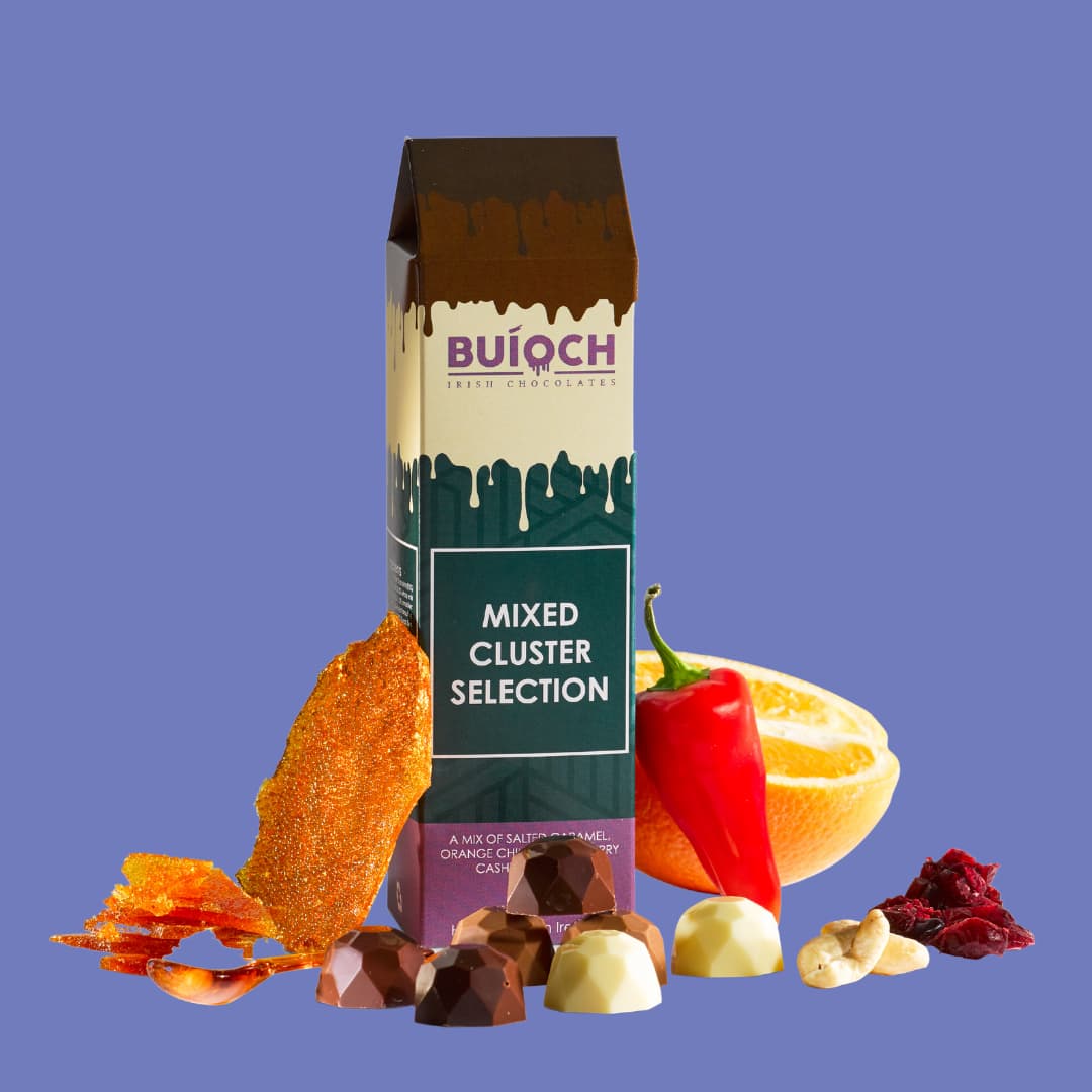 Mixed cluster selection. Milk chocolate salted caramel clusters, white chocolate cranberry cashew clusters and dark chocolate orange and chilli clusters. Handamde by Buíoch Irish Chocolates. Packaging, clusters and ingredients on a blue background