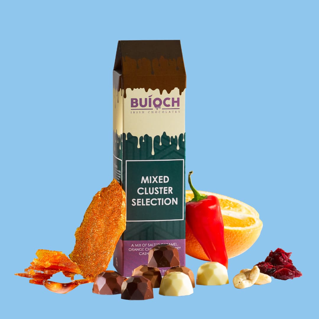 Mixed cluster selection. Milk chocolate salted caramel clusters, white chocolate cranberry cashew clusters and dark chocolate orange and chilli clusters. Handamde by Buíoch Irish Chocolates. Packaging, clusters and ingredients on a blue background