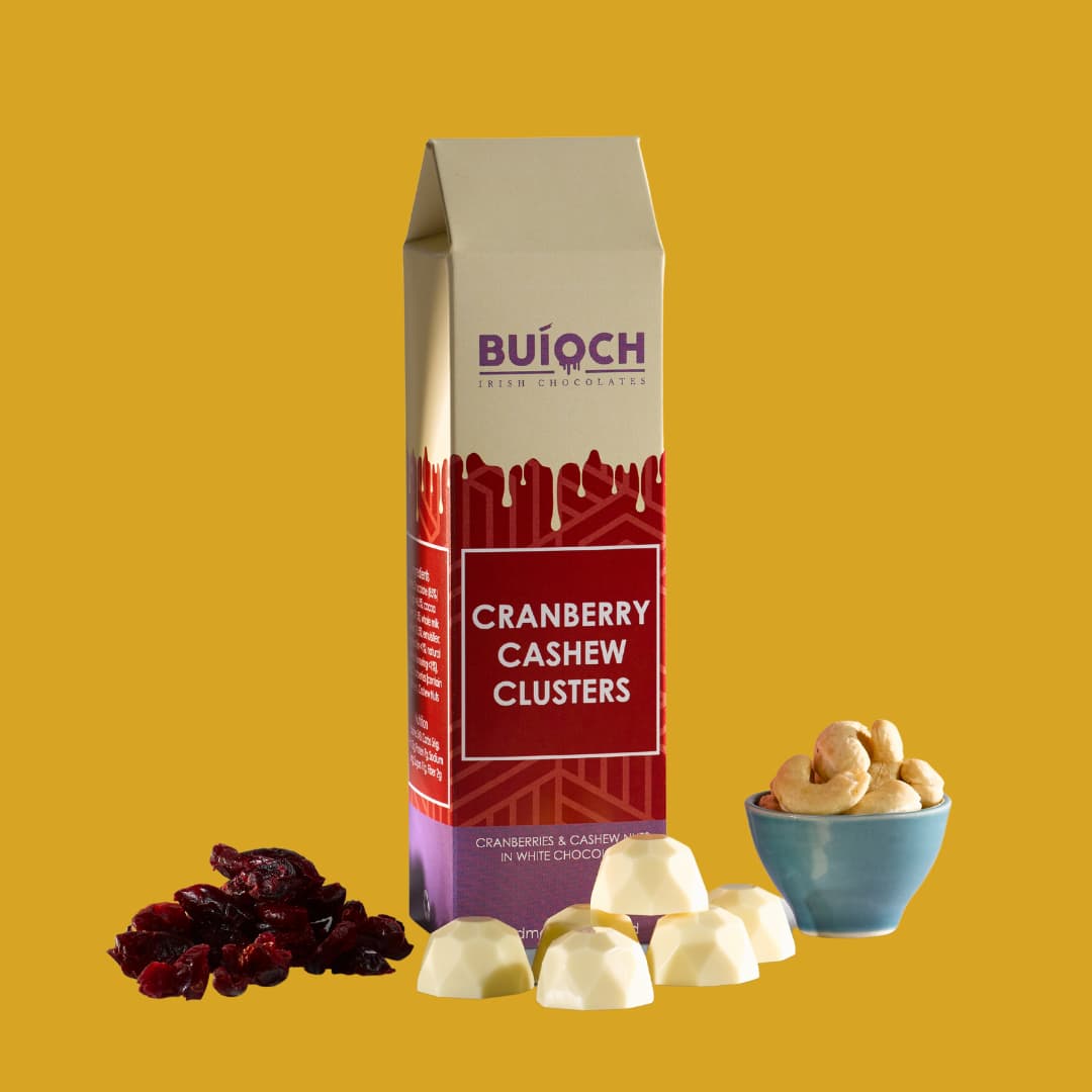 Crenberry Cashew Clusters - Cranberries and Cashew Nuts in White Chocolate. Handamde by Buíoch Irish Chocolates. Packaging, clusters and ingredients on a gold background