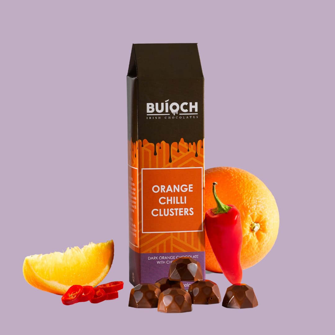 
                  
                    Orange Chilli Clusters - Dark orange chocolate with chilli flakes. Handamde by Buíoch Irish Chocolates. Packaging on a light purple background
                  
                
