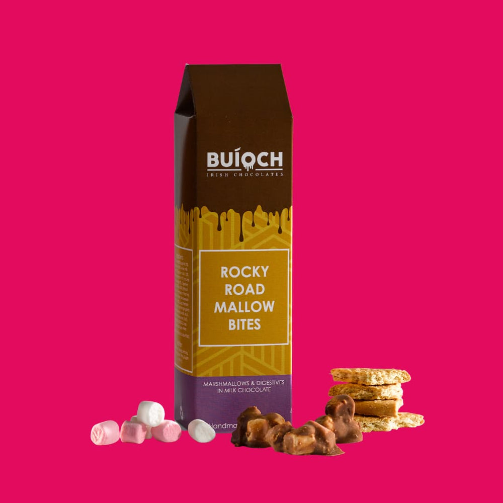 Rocky Road Mallow Bites - Marshmallow and Digestives in Milk Chocolate. Handmade by Buíoch Irish Chocolates. Packaging, Bites and Ingredients on a Pink Background.