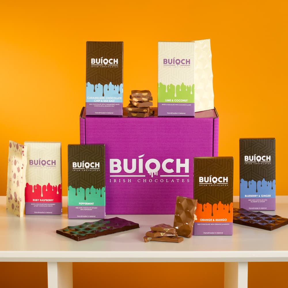 The Box O'Bars, six handmad Buíoch Irish chocoalte bars on a white table top surrounding of purple mailer box all against an orange background.
